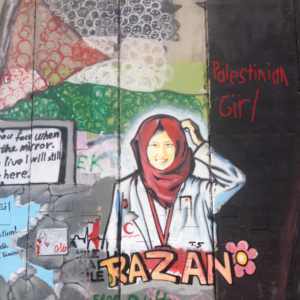 a wall of graffiti: centered is image of a woman, "RAZAN", a smiling woman in hijab; griffiti of Palestinian flag above her; "Palestinian Girl" written top right. cover art from "Bethlehem wall graffiti Razan with flower": "Portrait graffiti of 21-year-old nurse Razan al-Najjar with a flower by the Palestinian Artist Taghi Al Din on West Bank side of the Israeli West Bank barrier in Bethlehem from 2018. Razan al-Najjar was killed while she was doing her job at protests in front of the Gaza–Israel barrier on 1 June 2018."